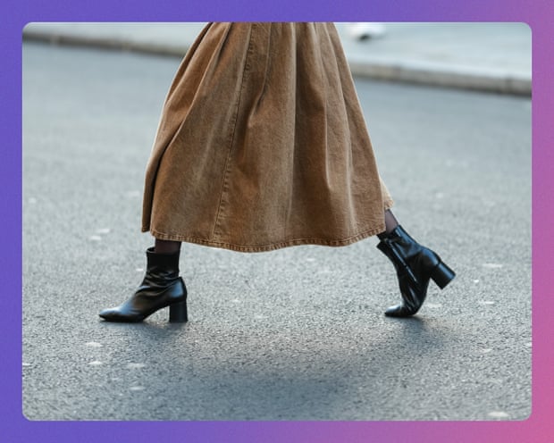 A person's legs in a long tan skirt and shiny black heeled ankle boots mid-stride