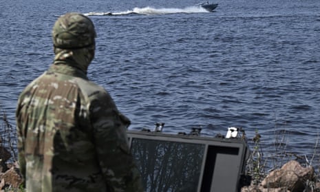 A Ukrainian serviceman, wearing a camouflage uniform, stands with his back to the camera, facing open water, as he controls a naval drone that is rippling the water in the distance. 