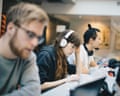 A woman with headphones rests her head on her hand, focused on a laptop, beside coworkers at a desk