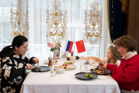 Two women and a young girl sit eating at a table with a white tablecloth and a small Russian flag and Chinese flag placed on it as decoration