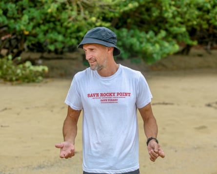 A man in a blue hat, white T-shirt emblazoned with Save Rocky Point standing on a beach