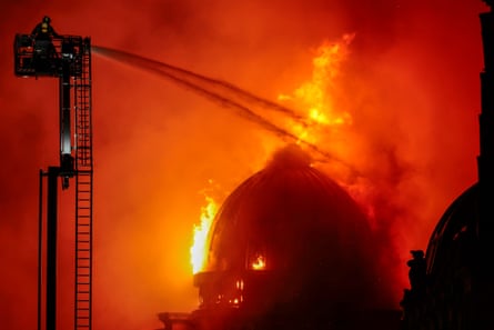 Firefighter on crane sprays hose on to building engulfed in flames.