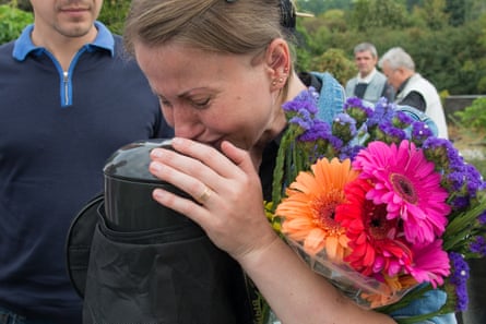 A woman holding a bunch of flowers cries while kissing an urn