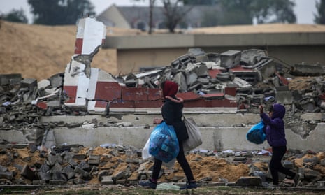 Two girls carrying plastic bags walk past a destroyed house
