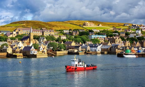 A red based boat sails in the foreground. Behind it is four rows of buildings build on hills; the tops of the hills are visible in the background.