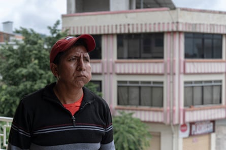 A middle-aged man from an Indigenous community frowns as he stands on the balcony of a building. His cheek is bulging, perhaps from chewing coca leaves 