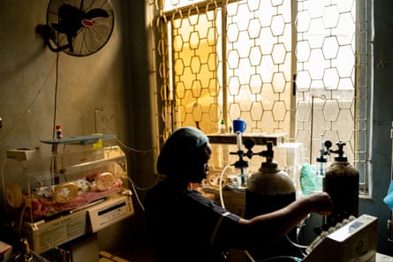 An African nurse in a dark room checks oxygen levels as she tends to a tiny baby in an incubator