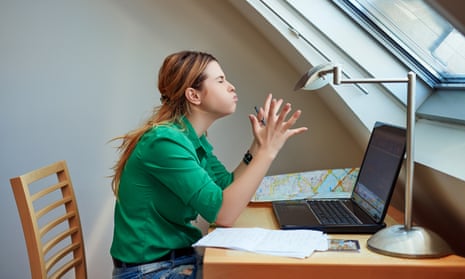 Angry young white woman sitting at a desk. She is wearing a green shirt and jeans and is stretching out her hands and scrunching her eyes shut in frustration.