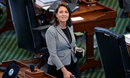 A picture taken from above of a middle-aged white woman with long brown hair, a gray suit coat and black skirt, holding a glass of coffee as she walks across a green carpet behind two tables, looking directly into the camera and smiling.