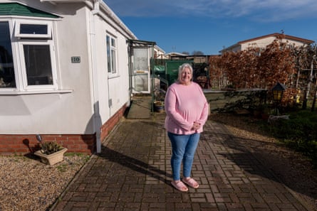 she has grey-blond hair in a ponytail and wears a pink sweatshirt and sliders with blue jeans.