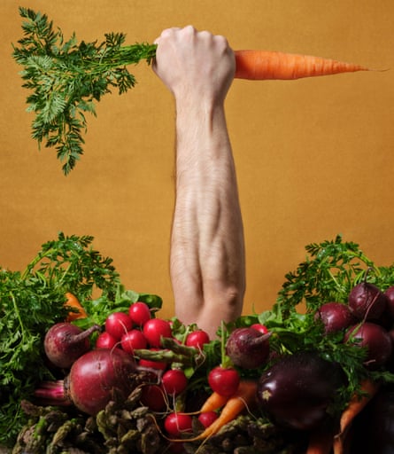 Man’s arm, with fist holding a carrot, sticking up through a pile of fruit and veg