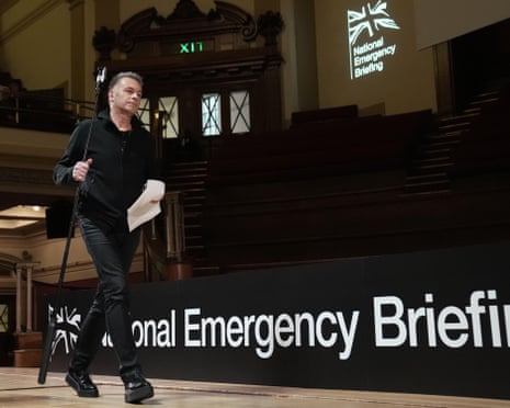 A man dressed in a black sweatshirt and jeans carrying a microphone stand walks off stage. In the backdrop is a the logo of the National Emergency Briefing with a British flag on it
