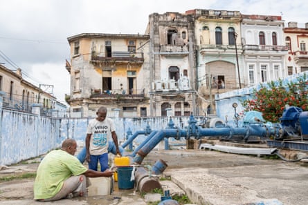 Two men fill containers with water at a pumping station in the centre of Havana, with dilapidated buildings in the background.