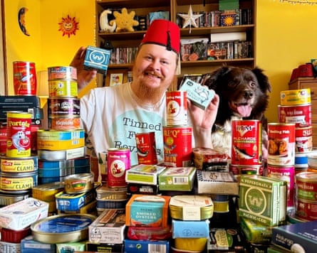 A man sitting at a table alongside a dog, with piles of tinned fish on the table