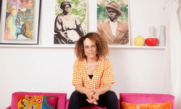 Bernardine Evaristo, photographed at her home: she wears black trousers and an orange and white checked shirt, and is sitting on a bright pink sofa with orange and patterned cushions in front of a white wall which has, behind her head, a shelf with two tall white vases and two smaller vases, one red and one yellow. There are three pictures on the wall; two are tinted vintage photographic portraits of Black people, one female, and one male.