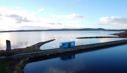 Blue mobile library van drives over a narrow stone causeway across an expanse of water; the cloudy blue sky is reflected in the water
