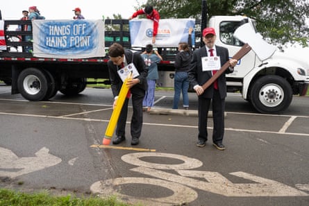 Two men in suits hold an oversized axe and pencil as people on a truck in background hold banner reading ‘hands off Hains Point’