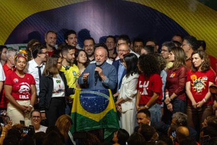 Lula speaks into a microphone holding a Brazilian flag, surrounded by supporters on stage