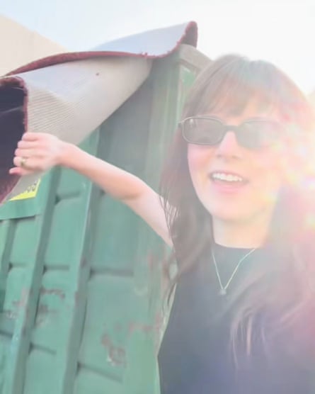 A woman pulls a piece of red carpet out of a green skip.