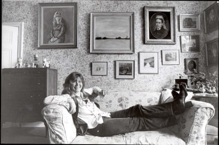 Black and white pic of Jilly Cooper sitting on a sofa in a drawing room with framed pictures on the wall and patterned wallpaper.