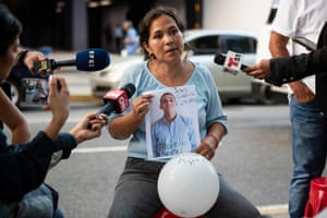 A woman holds a A4-sized photograph of a young man while surrounded by reporters holding microphones