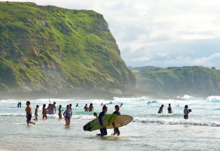 Surfers walking in the surf carrying surfboards along a beach with a large grassy cliff in the background