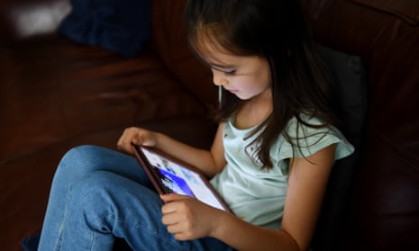A young girl with long brown hair is sitting on the floor with an iPad resting on her bent knees. She is wearing jeans and a light green top and is looking at the screen