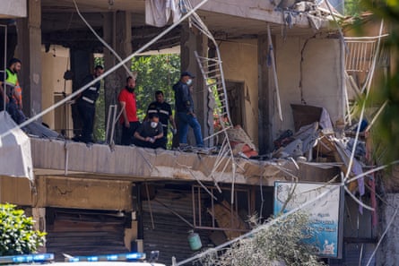 Police officers and emergency personnel stand and crouch on a floor of an apartment that has had its walls blown off.