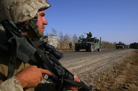 A man holding a machine gun stands by the side of a dirt road on which armoured vehicles are travelling