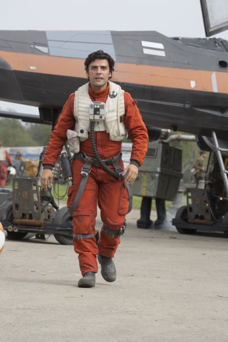 Oscar Isaac as Poe Dameron, wearing an orange jumpsuit and white vest with tubes and harnesses attached, walking away from a fighter plane towards the camera.