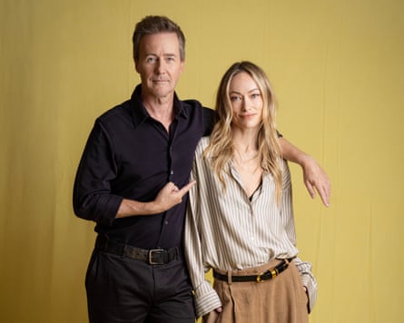 Ed Norton has one arm over Olivia Wilde’s shoulder and is pointing at her with the other hand. She wears loose striped open-neck shirt and baggy trousers