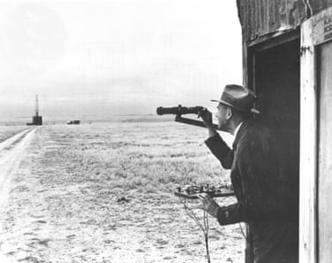 Dr. Robert H. Goddard observes the launch site from his launch control shack while standing by the firing control panel. From here he can fire, release, or stop testing if firing was unsatisfactory. Firing, releasing, and stop keys are shown on panel. The rocket is situated in the launch tower.