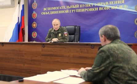 Vladimir Putin, dressed in military fatigues, speaks with Valery Gerasimov; Putin sits at a high desk with Gerasimov, also in fatigues, seated below with his back to the camera and paperwork laid out before him.