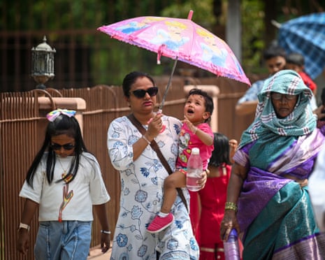 A woman shields a toddler with a pink umbrella while walking outdoors on a hot day