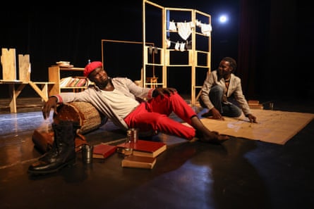 An African man in a red beret reclines against a cushion as another African man squats over a map on a sparsely furnished stage