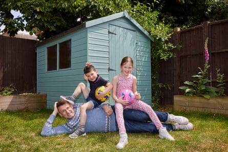 Tim Jonze with his children. They are in a garden in front of a shed. Tim is lying on his side with his children are sitting on him.