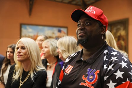 A man in a Maga hat and stars and strips jacket stands in a room surrounded by blond women.