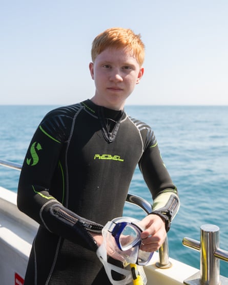 A boy with red hair on a boat at sea wearing a wetsuit and holding a snorkel and mask, with other young people behind him.