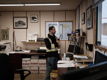 Truck driver Andy Rae checks his radio in an office; he wears a yellow hi-vis vest and black hoodie.