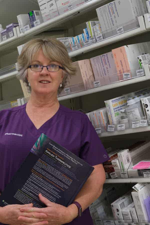 Kate Wood, in her pharmacist uniform, stands in front of shelves of medicine.