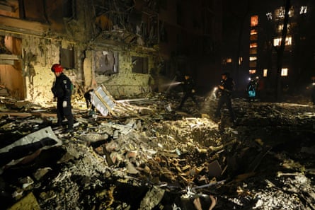 Rescue workers walk in front of a damaged residential building with piles of rubble in the street.