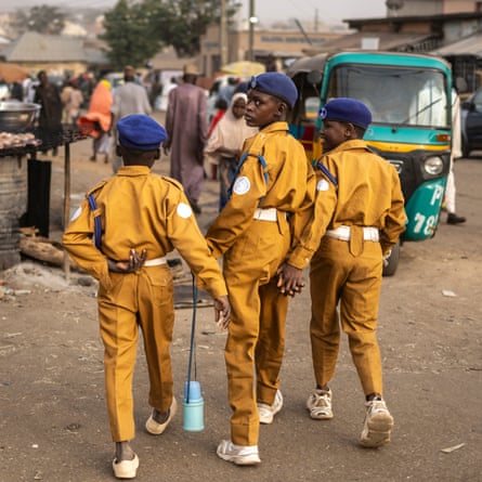 Three boys wear mustard-yellow jumpsuits with white belts, trainers, and dark blue berets; one carries a blue thermos flask.