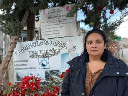 A young Indigenous woman by a sign in Spanish under pine trees that says ‘Colla community – Finca el Chañar.’