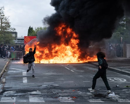 Two young people run across a road as a fire burns in the background