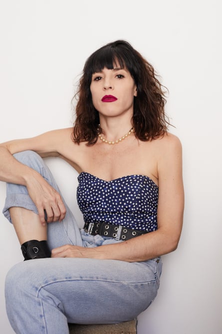 Marina Otero with shoulder-length dark wavy hair, wearing a navy blue and white polka dot bustier top and jeans, sitting with one knee up and her arm resting on it, staring at the camera with intensity.