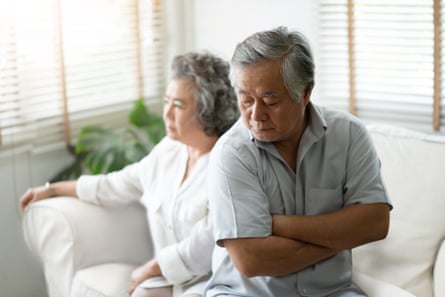 Unhappy Asian senior man sitting on the edge of the sofa with arms crossed, with his wife sitting behind him, looking in the distance.