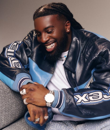 A smiling man with a beard sits wearing a black and blue leather jacket with jewelry