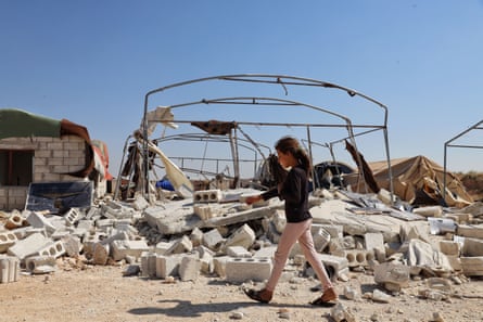 A girl walks past rubble and the frame of a tent