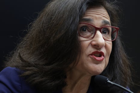 Middle-aged white woman with thick dark hair and red-framed glasses, wearing red lipstick, speaks into a microphone.
