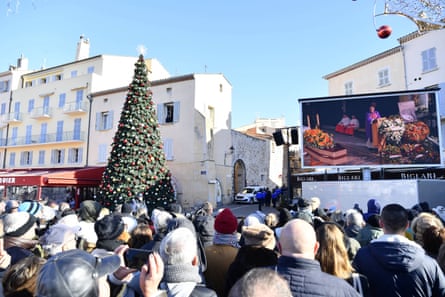 People gather in front of a giant screen showing the ceremony in a public square; there is a tall Christmas tree to one side and the square is lined with tall, pale yellow buildings.